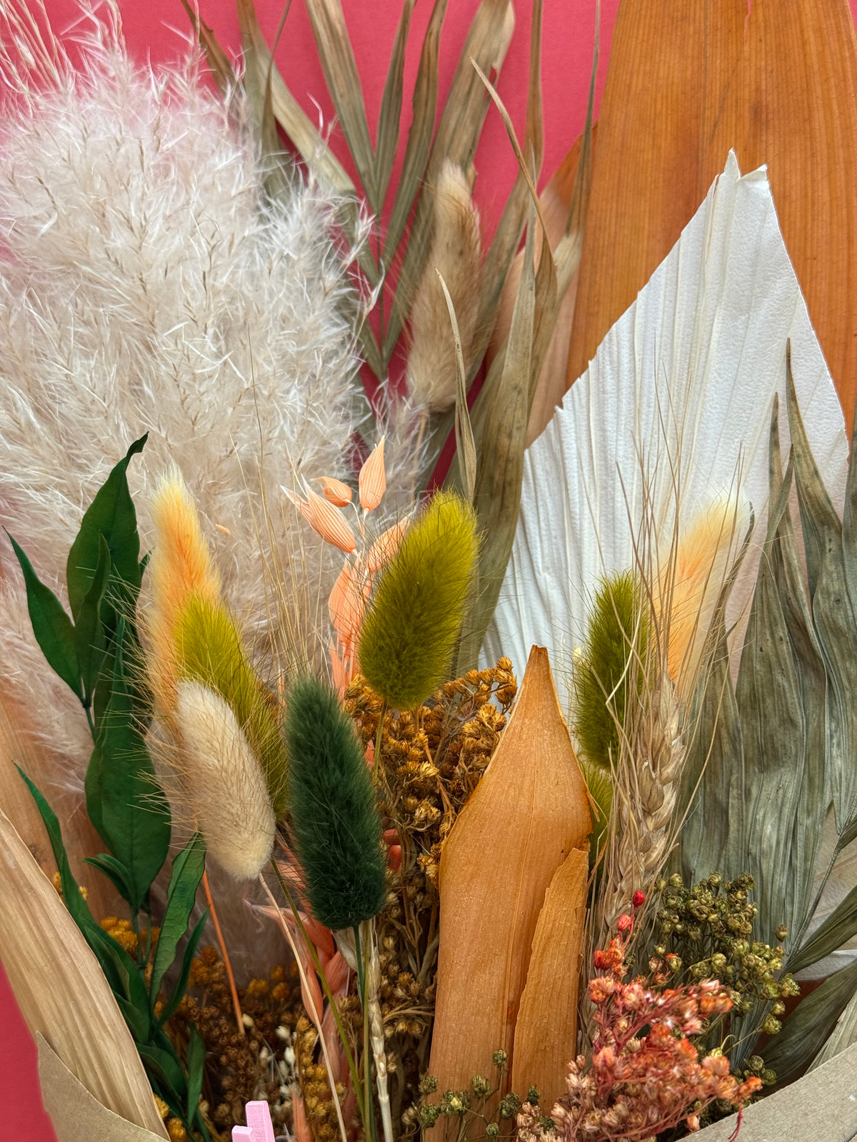 A close-up of a dried floral arrangement featuring fluffy white pampas grass, green and yellow bunny tail stems, tan leaves, and small pinkish flowers against a pink and wood background.