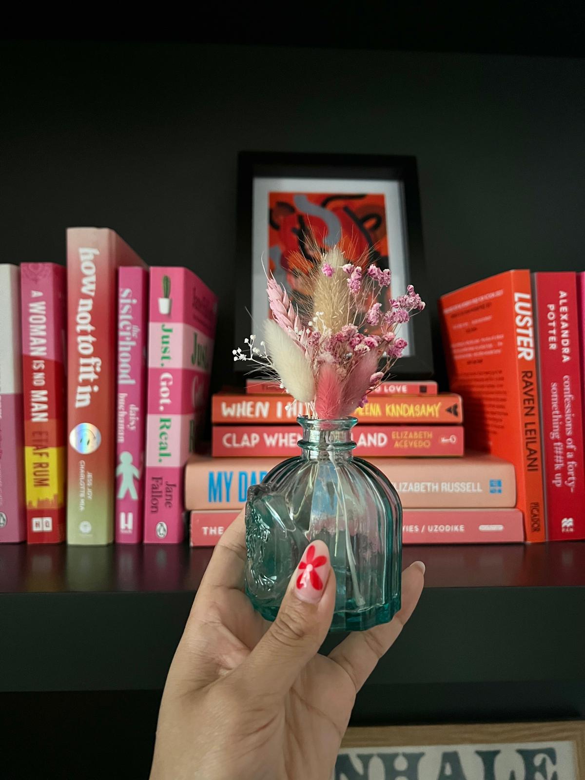 Hand holding a decorative bottle with flowers in front of a bookshelf
