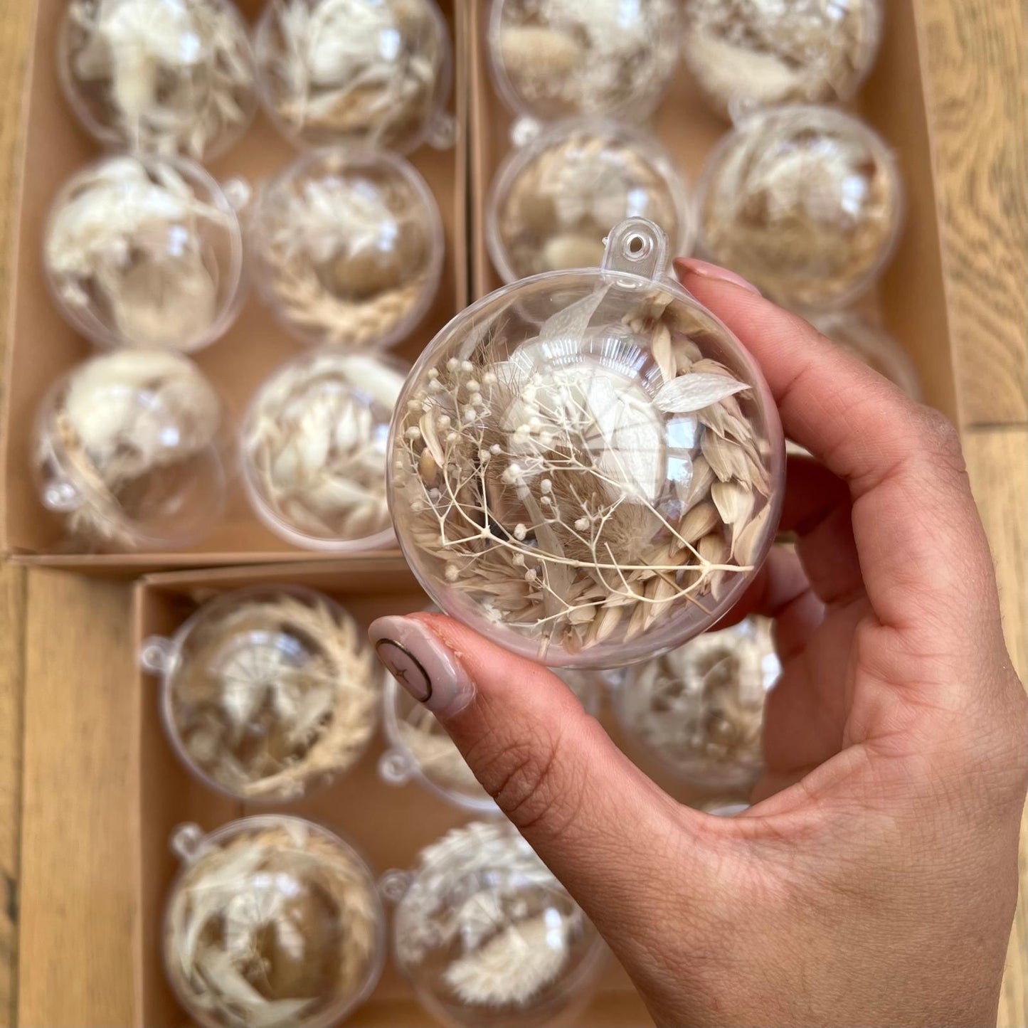 Hand holding a clear ornament with dried flowers against a background of similar ornaments in a box.