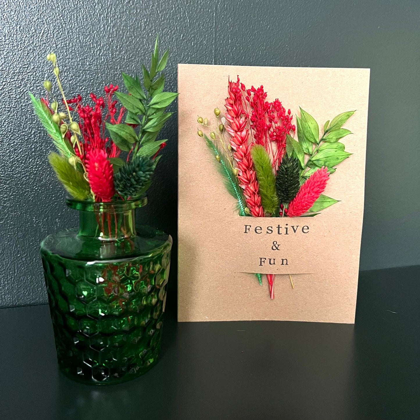 Green glass vase with red and green floral arrangement next to a Christmas card with floral design and 'Festive & Fun' text.