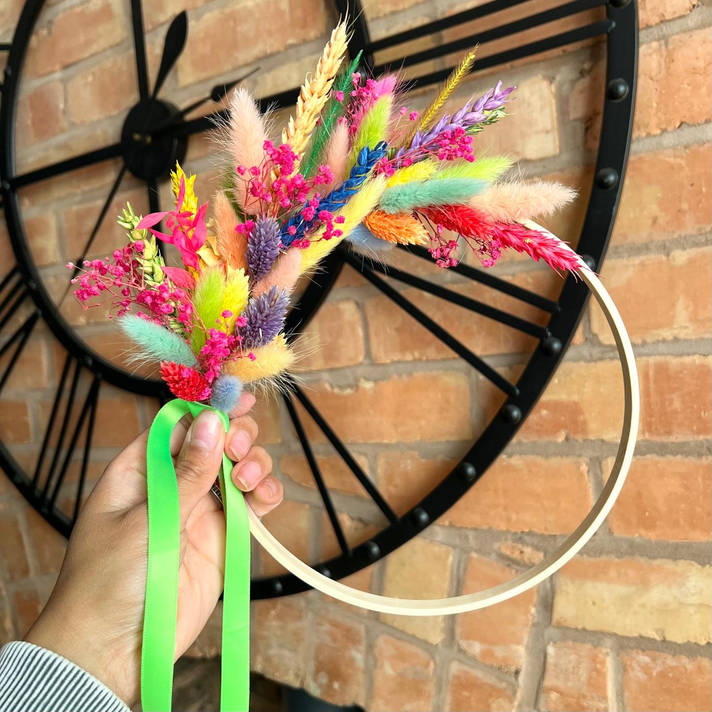 Hand holding a handmade dried flower wreath with bold colours and green ribbon, styled against a brick wall and a clock