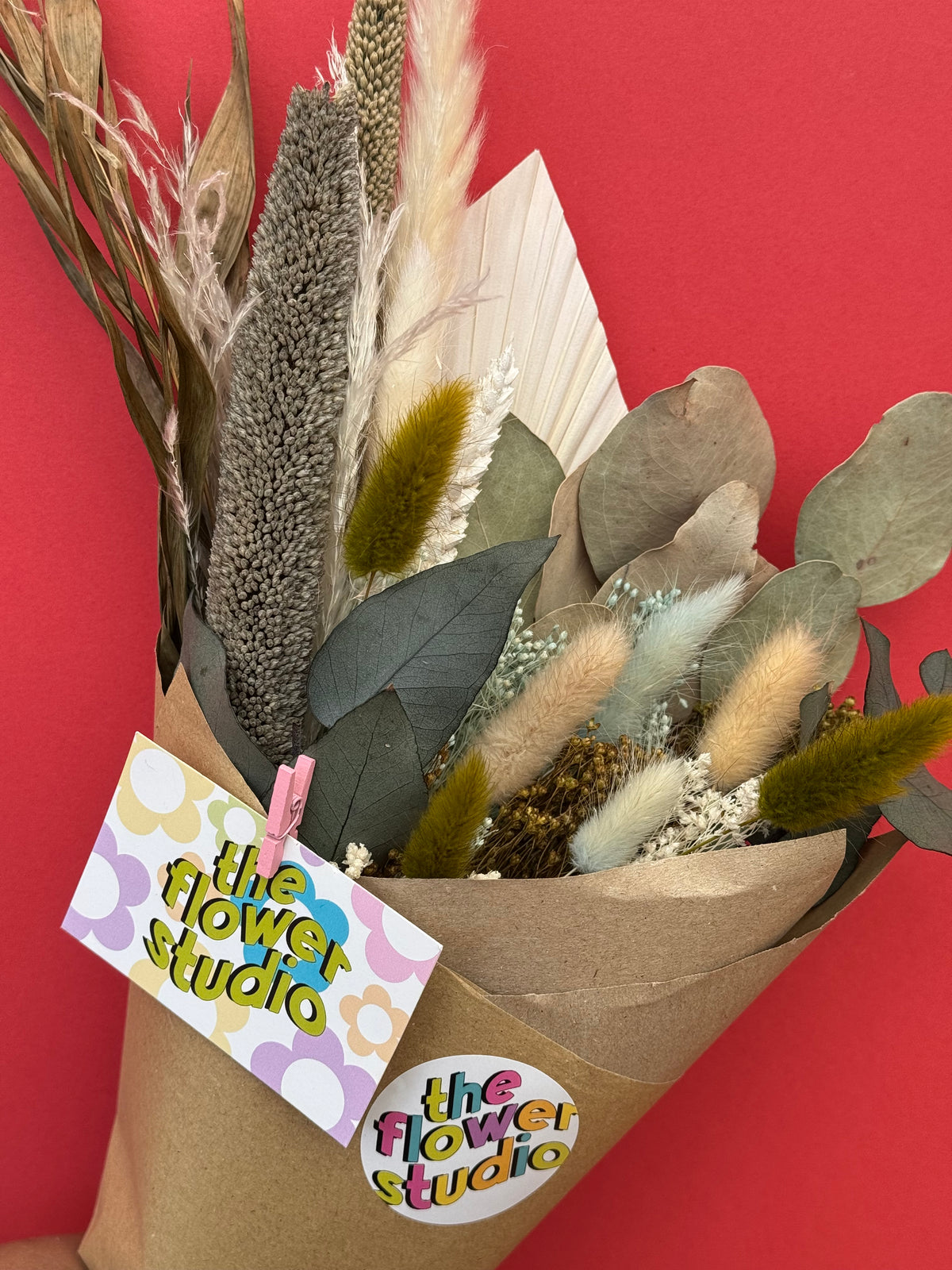 A dried flower bouquet wrapped in brown kraft paper with a paper tag reading 'the flower studio' against a solid red background.