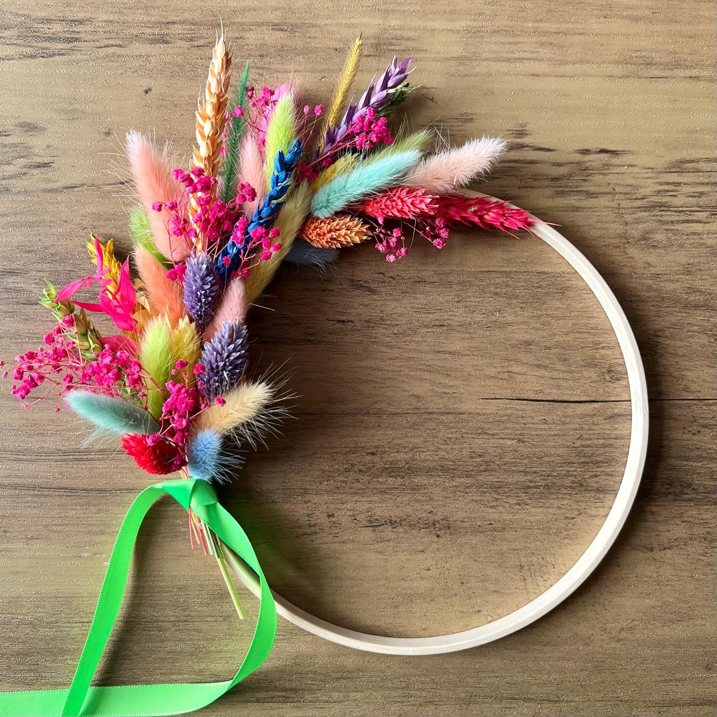 Bright rainbow dried flower hoop with bunny tails, grasses, and preserved blooms on a wooden ring laid on a wooden table.