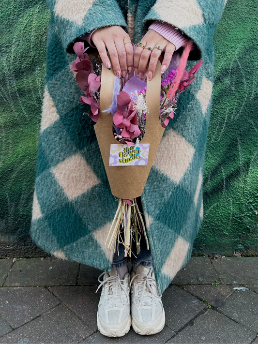 A person wearing a cosy green and cream checkered coat holds a bouquet of pink and purple dried flowers in a brown paper bag with a label that reads "The Flower Studio." Their hands, adorned with rings, gently grasp the bag handles. They stand against a textured green wall, with their feet clad in white sneakers, creating a stylish and artistic scene.