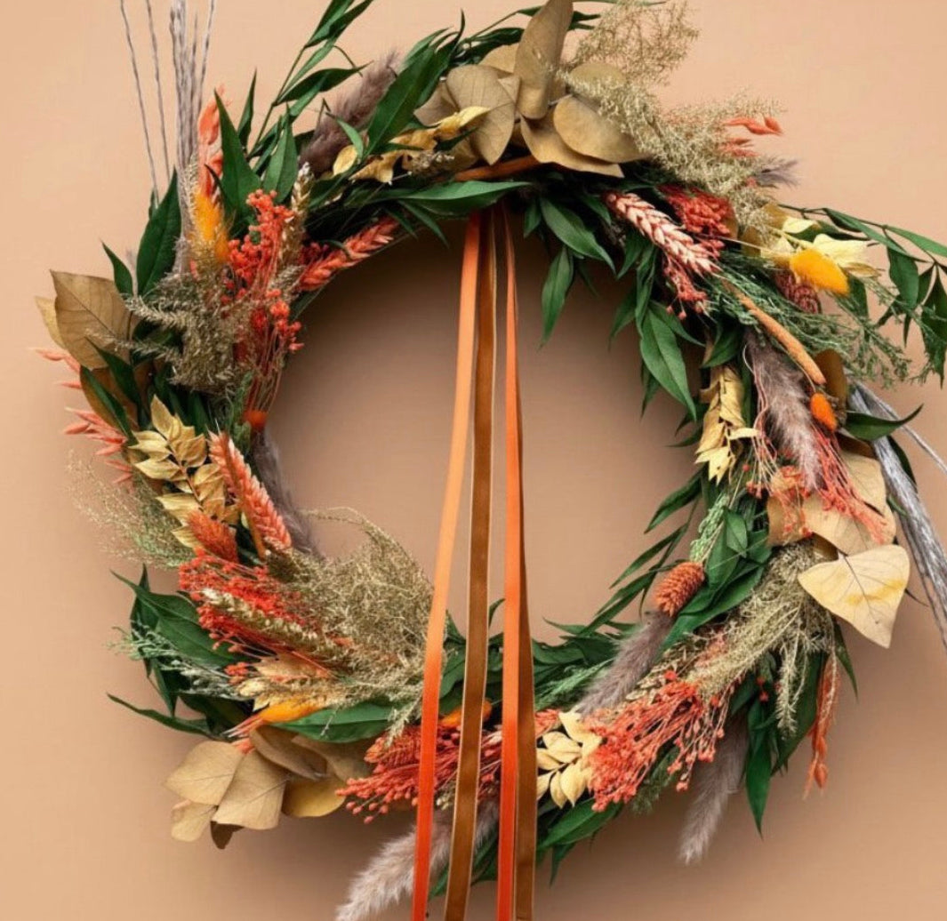 An autumn-themed dried flower wreath with green leaves, orange and beige botanicals, and two long orange-brown hanging ribbons, displayed against a solid warm beige background.i