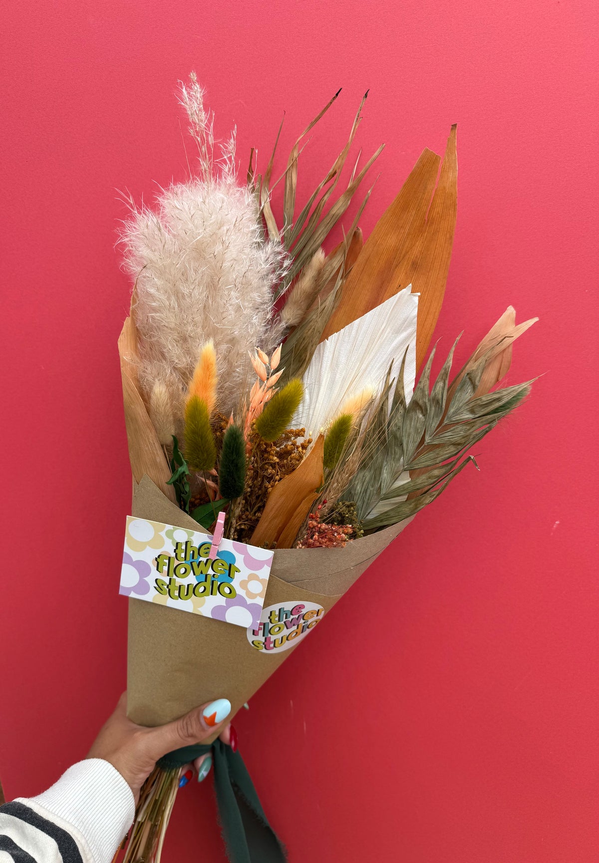 A hand holding a dried flower bouquet wrapped in brown kraft paper, featuring pampas grass, orange leaves, and green foliage, against a solid red background. The bouquet has a paper tag reading 'the flower studio'.