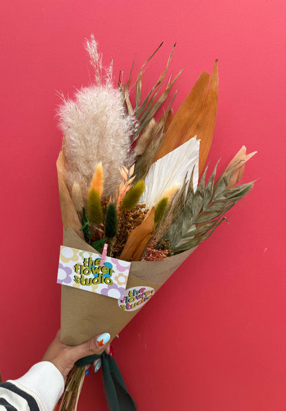 A hand holding a dried flower bouquet wrapped in brown kraft paper, featuring pampas grass, orange leaves, and green foliage, against a solid red background. The bouquet has a paper tag reading 'the flower studio'.