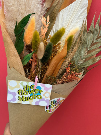 A dried flower bouquet wrapped in brown kraft paper, featuring various natural elements like pampas grass, wheat, and green leaves, with a 'the flower studio' tag attached, set against a solid red background.