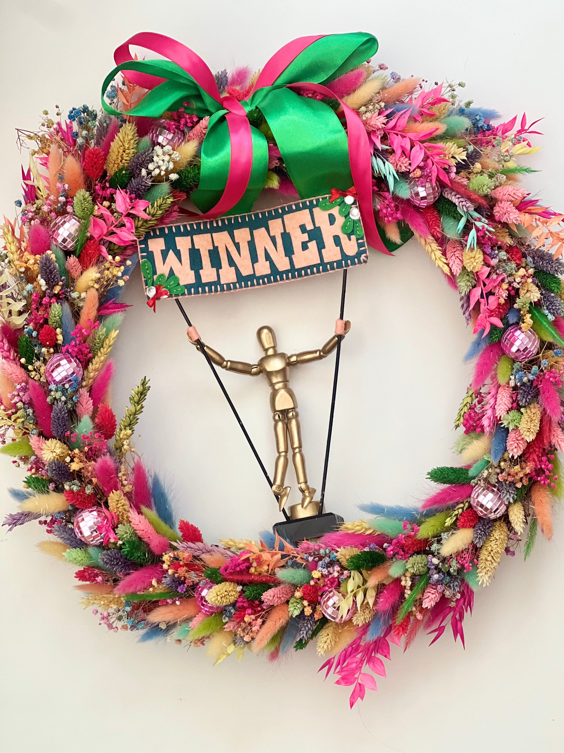 An award-winning rainbow wreath featuring vibrant colours, including red, yellow, blue, green, purple, orange, and various shades of these tones. At the centre of the wreath is a golden trophy, symbolising its win on Kirstie’s Handmade Christmas, as seen on Channel 4.
