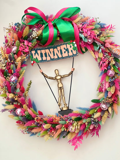 An award-winning rainbow wreath featuring vibrant colours, including red, yellow, blue, green, purple, orange, and various shades of these tones. At the centre of the wreath is a golden trophy, symbolising its win on Kirstie’s Handmade Christmas, as seen on Channel 4.