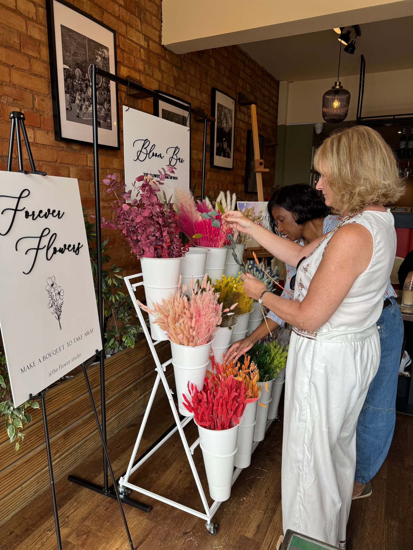 Two women arranging colorful dried flowers in white paper tubes on a display stand inside a room with wood floors and framed art.