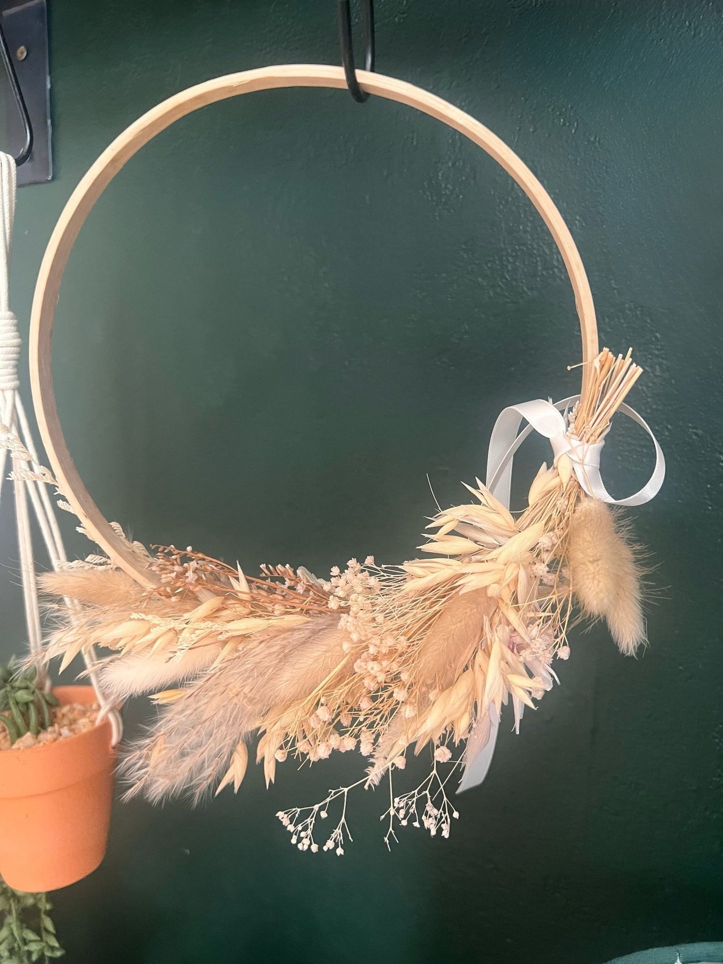 Rustic boho wreath made with soft dried flowers and natural textures, displayed against a green wall next to a small plant pot