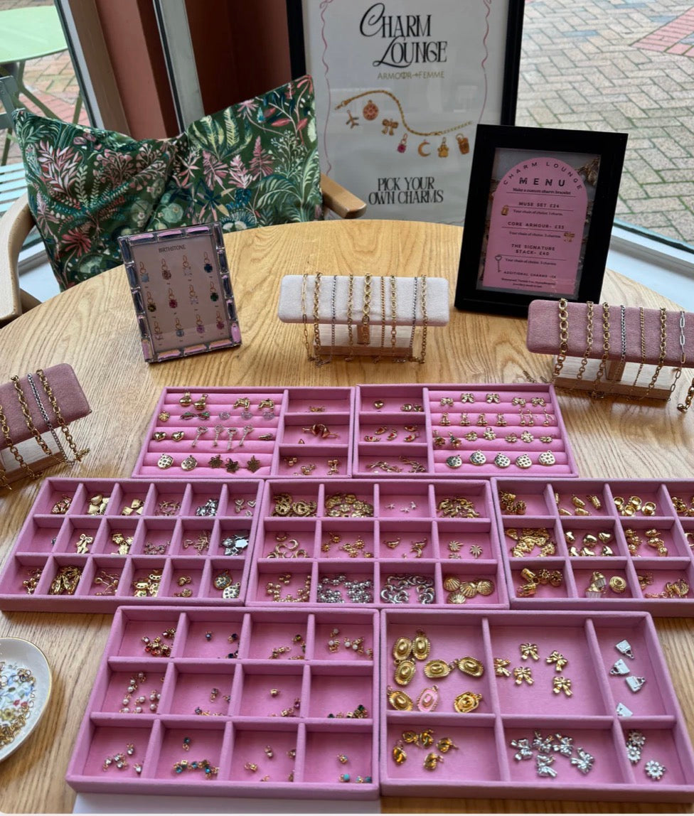 Pink jewelry organizer with various charms on a table, surrounded by jewelry display stands and a 'Charm Louise' sign.