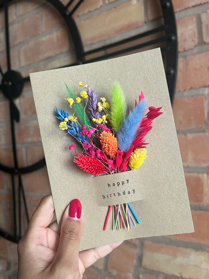  A hand holding a kraft paper birthday card decorated with a vibrant bouquet of dried flowers in rainbow colours, including yellow, red, blue, green, and purple. The card features the words “happy birthday” stamped in black lowercase letters below the bouquet, which is tied with multicoloured stems. A large black wall clock and brick wall are visible in the background.
