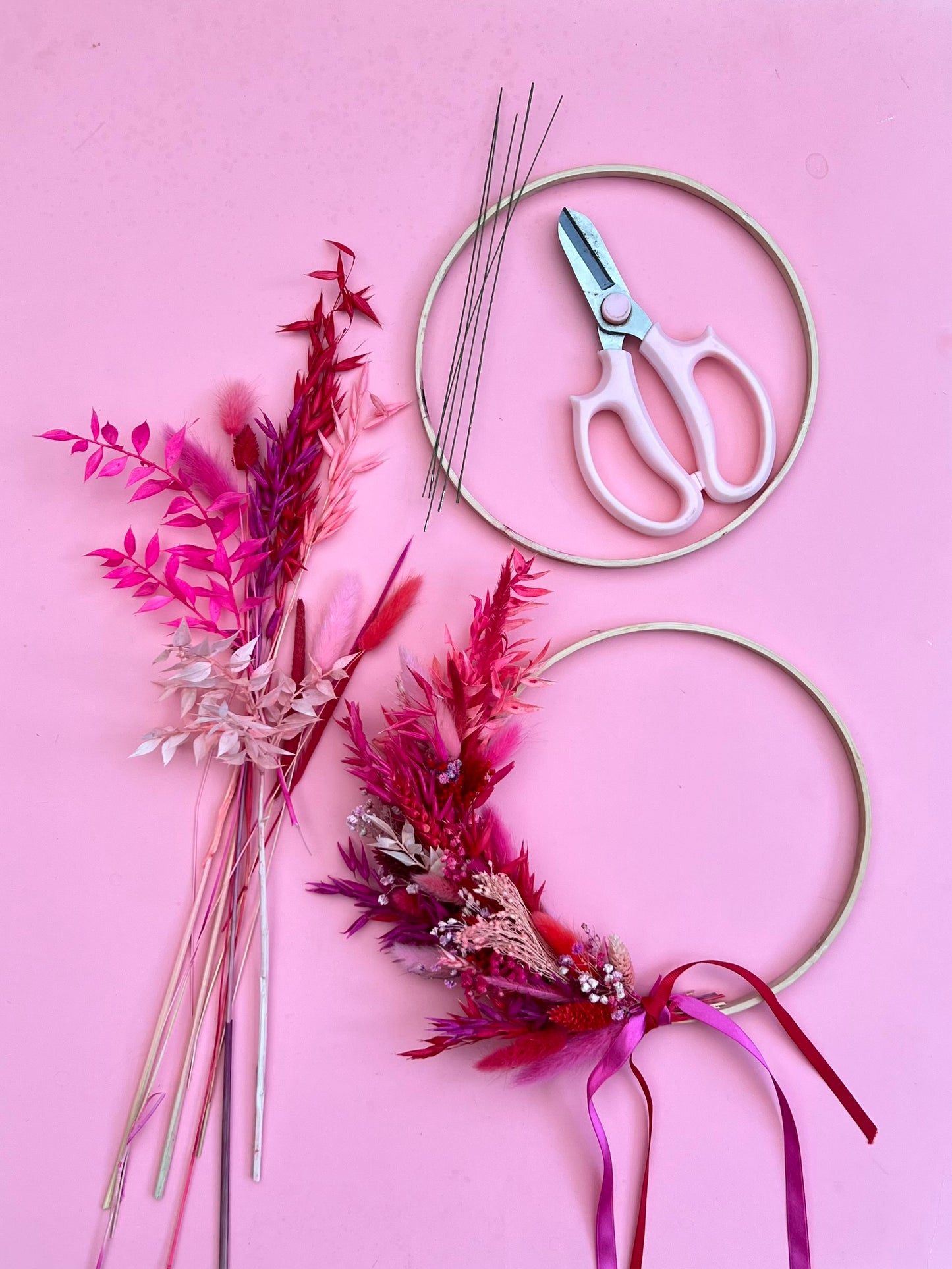 Decorative hoop with dried flowers, scissors, and ribbons on a pink background