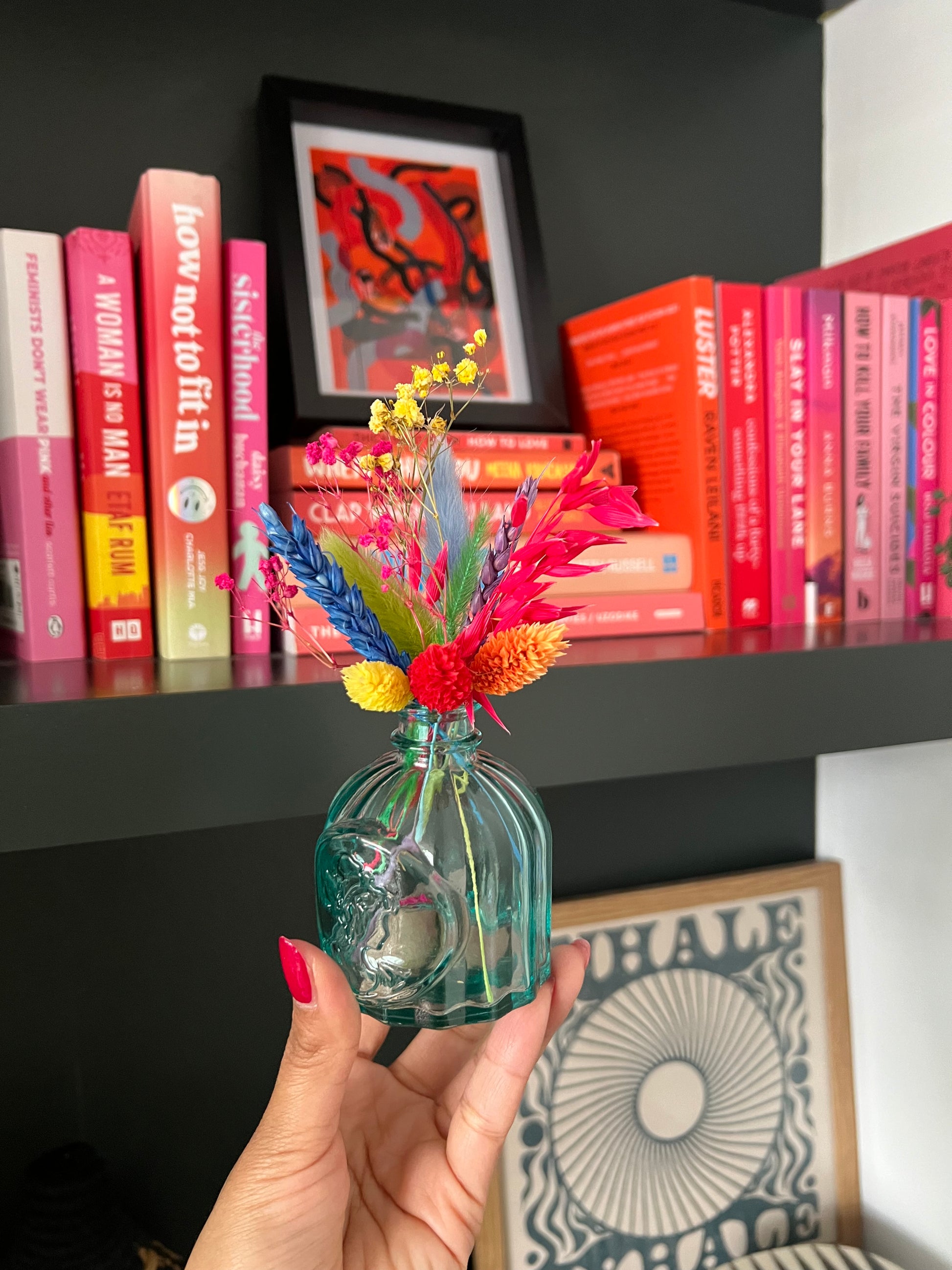  A hand holding a small teal glass vase filled with a vibrant arrangement of dried flowers in red, yellow, blue, green, and purple tones. The vase features a raised design of a woman’s profile. In the background is a bookshelf filled with pink and red books, a framed abstract red artwork, and a "WHALE" art print resting on the lower shelf.