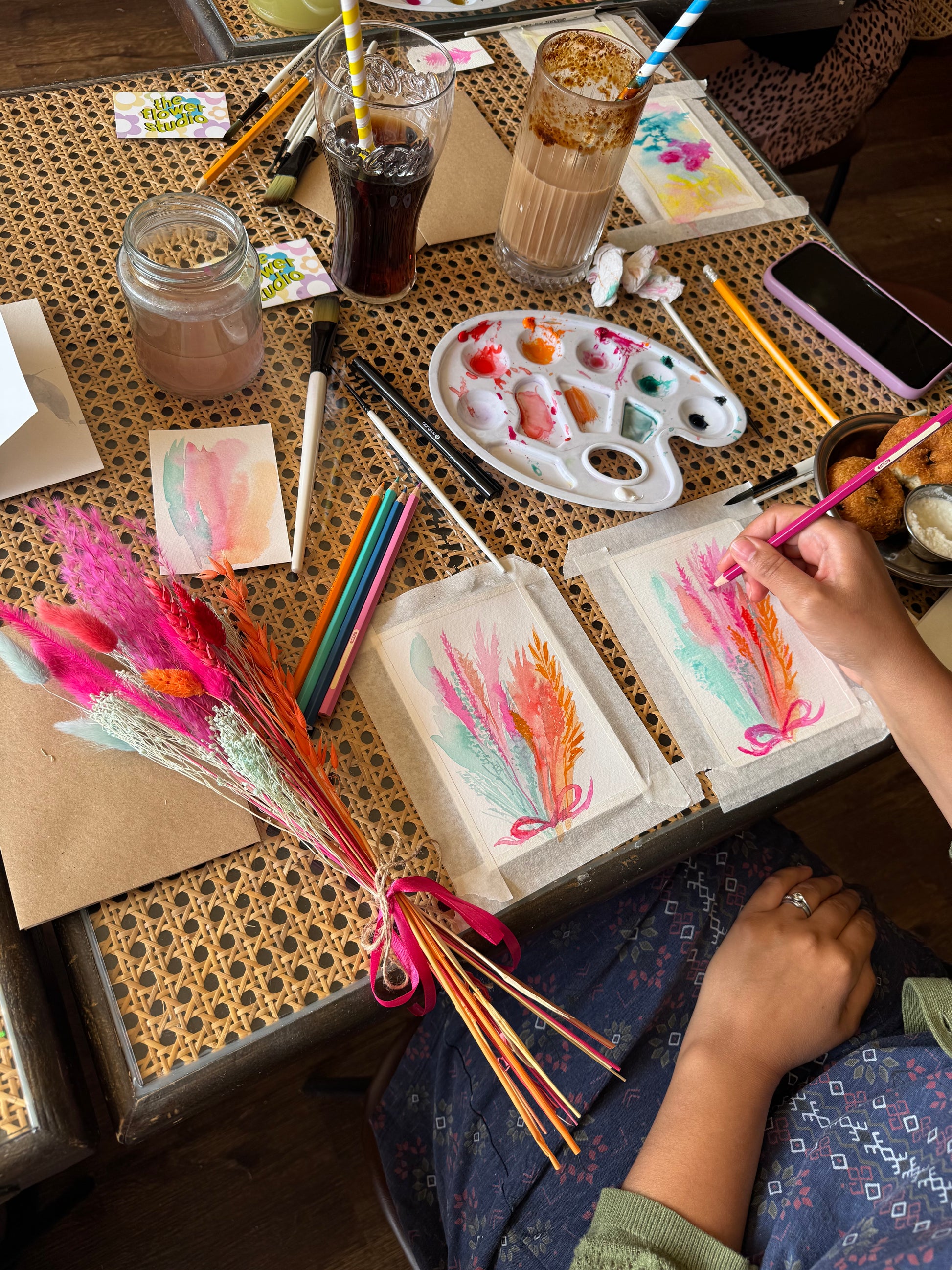Overhead view of a person painting two small art cards on a patterned table, surrounded by paintbrushes, a paint palette, pencils, mugs, and a bundle of colorful dried grass stems.