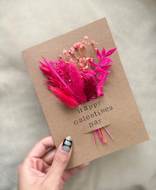 Card with pink flowers and feathers held by a hand, with 'Happy Galentine's Day' text.