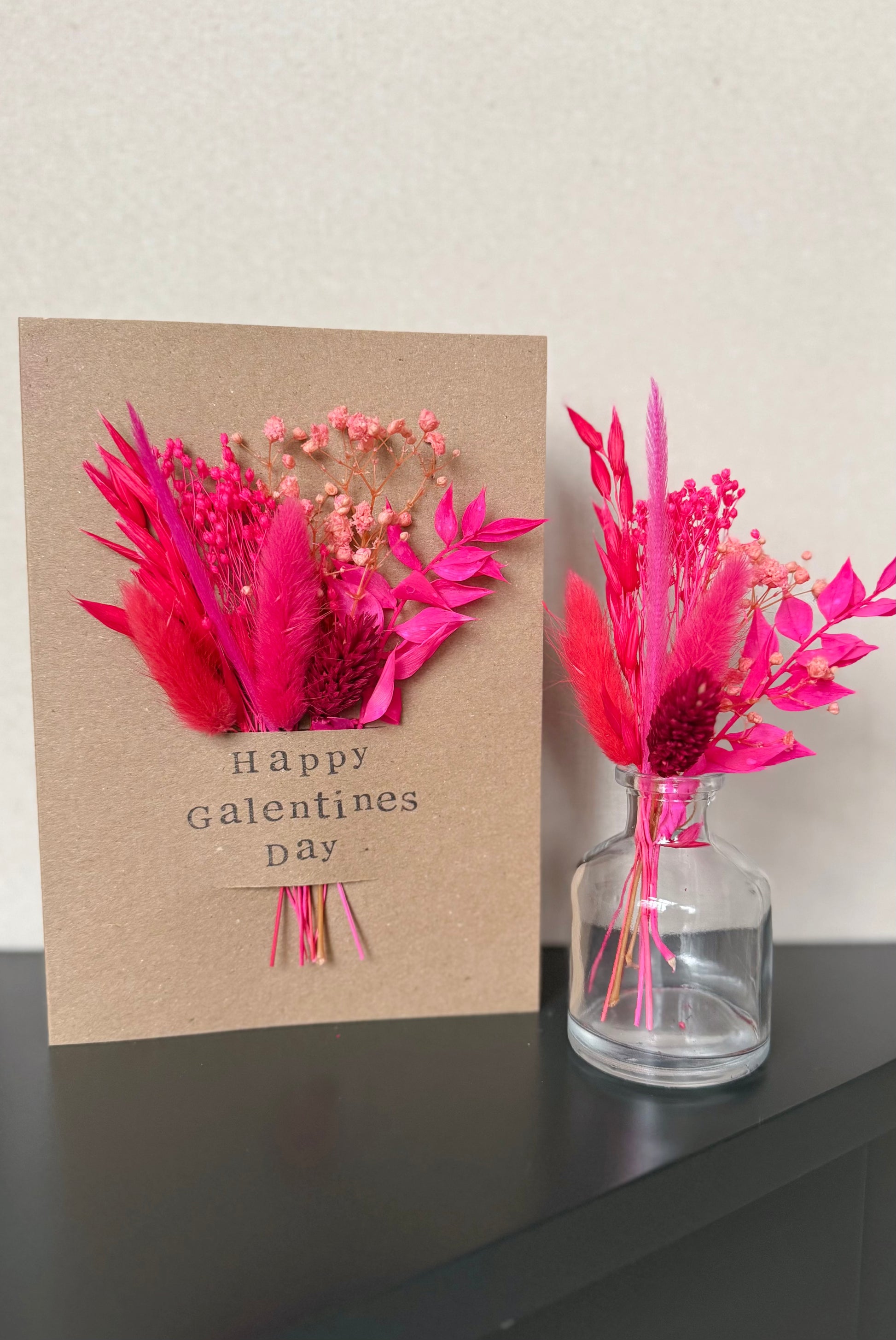 Card with pink flowers and 'Happy Galentine's Day' text next to a vase with similar flowers on a dark surface.