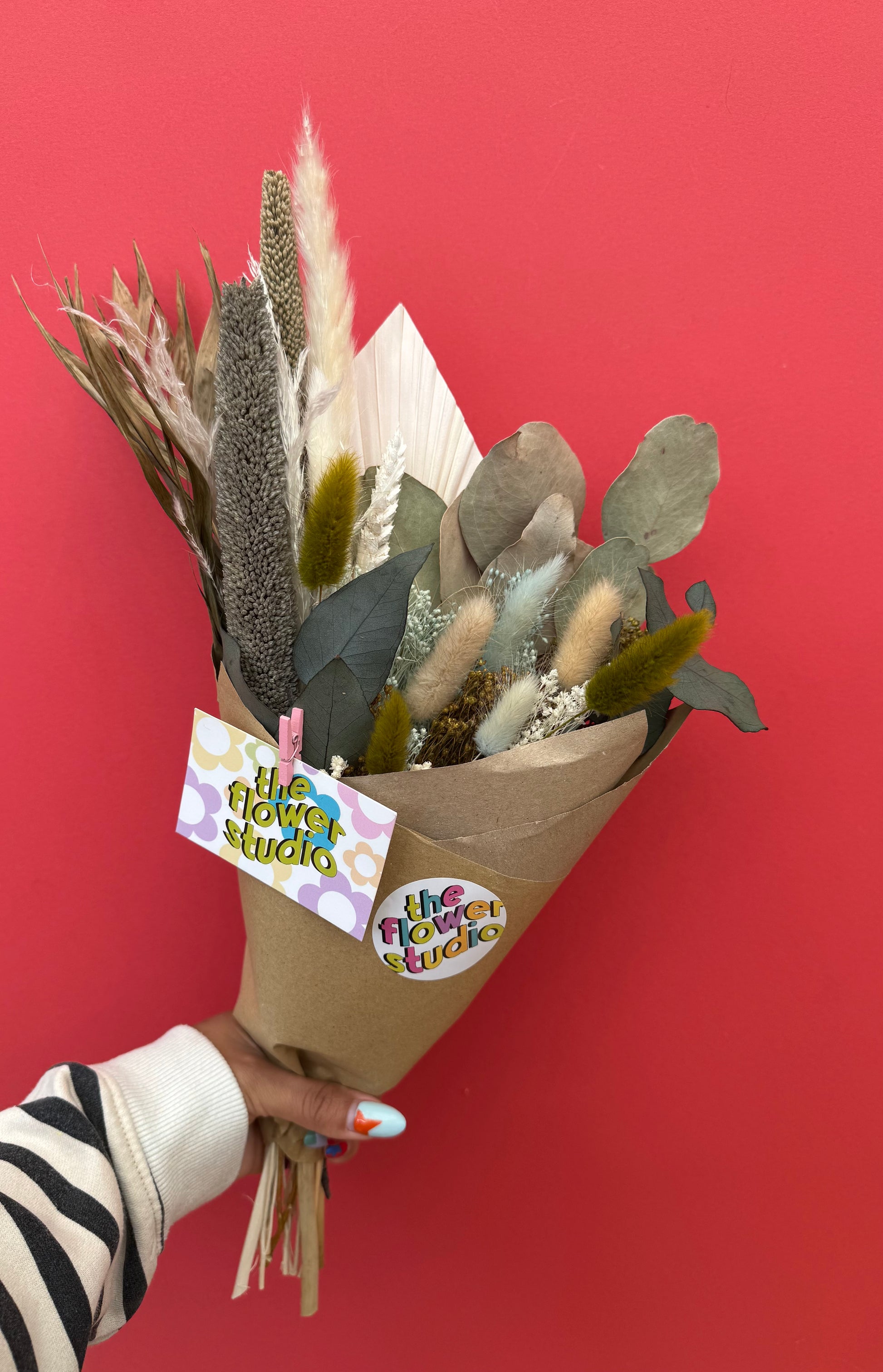 A hand holding a dried flower bouquet wrapped in brown kraft paper with a 'the flower studio' tag, set against a solid red background.