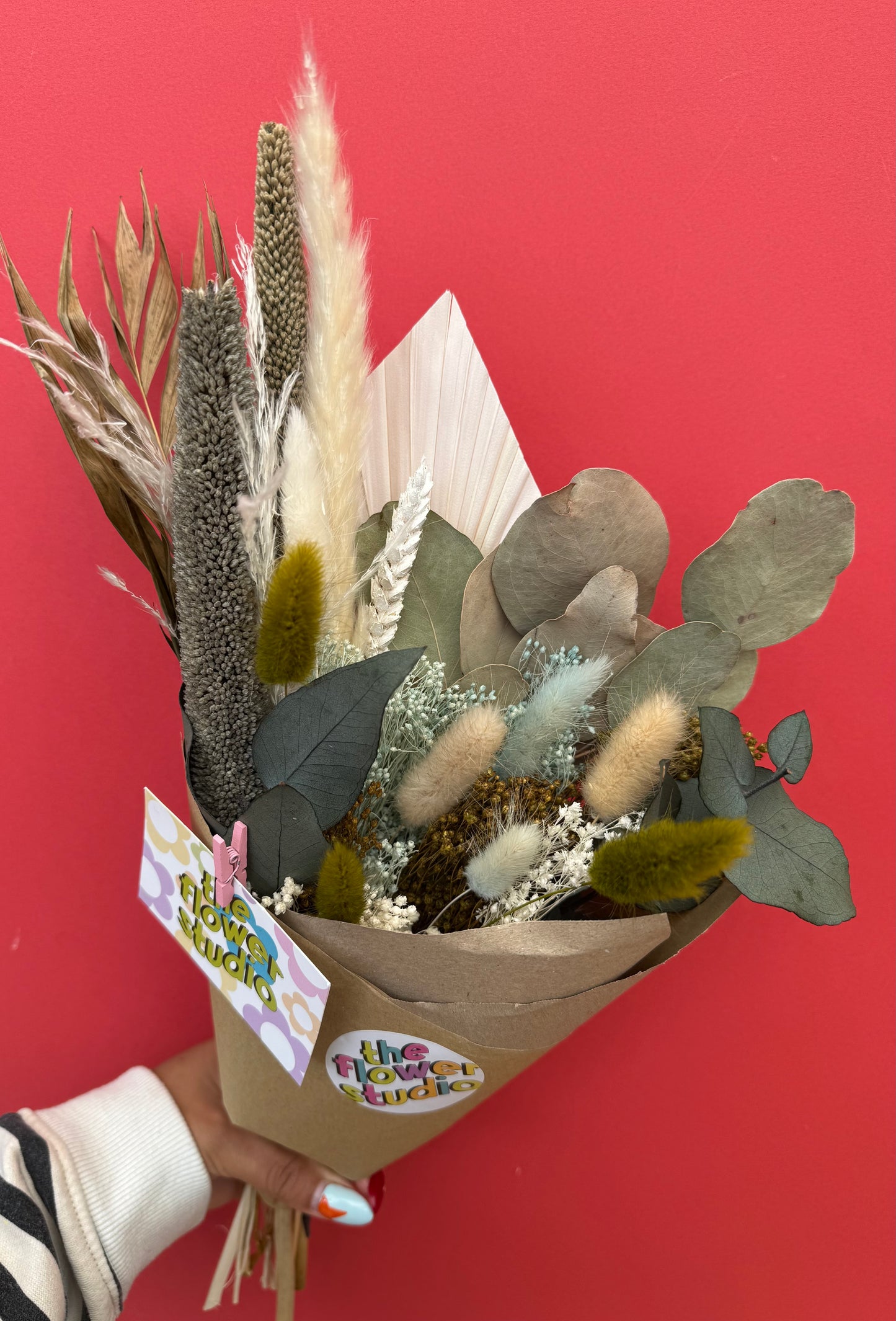 A dried flower bouquet held by a hand against a solid red background. The bouquet features natural dried grasses, bunny tails, eucalyptus leaves, and a small pale blue flower, wrapped in brown kraft paper with a 'the flower bundle' sticker and a gift tag.