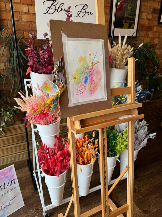 An overhead view of a decorative wooden display stand or easel holding multiple white cylindrical containers filled with colorful dried botanicals and flowers, with a small framed artwork resting on a cardboard support in the center.
