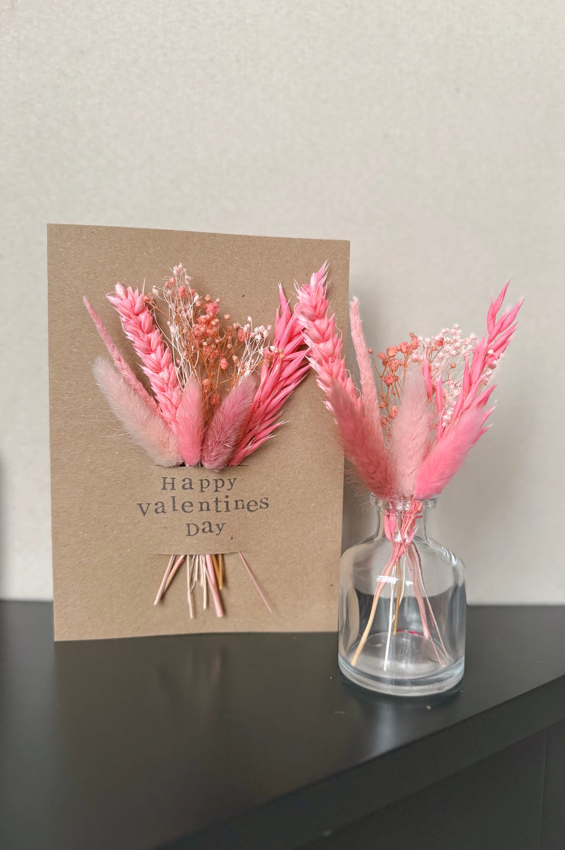 Valentine's Day card with pink flowers and a vase on a dark surface.
