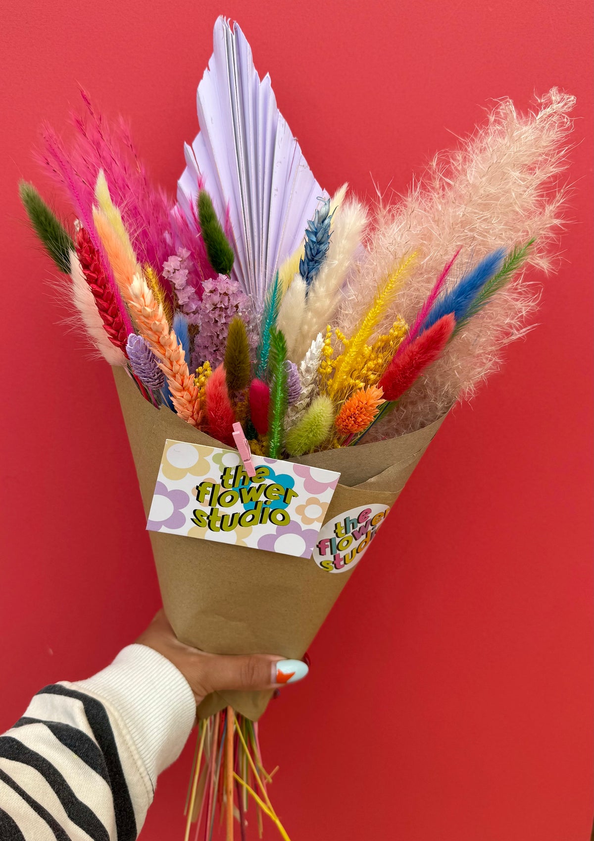 A hand holding a colorful dried flower bouquet wrapped in brown paper with a 'the flower studio' tag, set against a solid red background.
