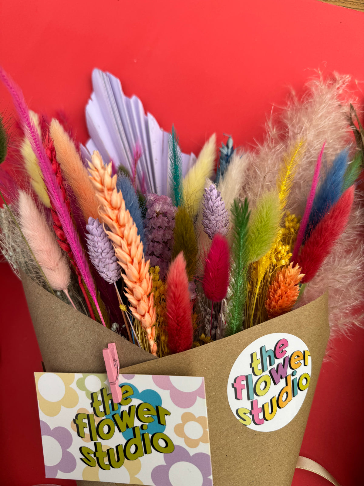 A colorful bouquet of dried decorative grasses and flowers wrapped in brown kraft paper, with a card and round sticker reading 'the flower studio' against a bright red background.