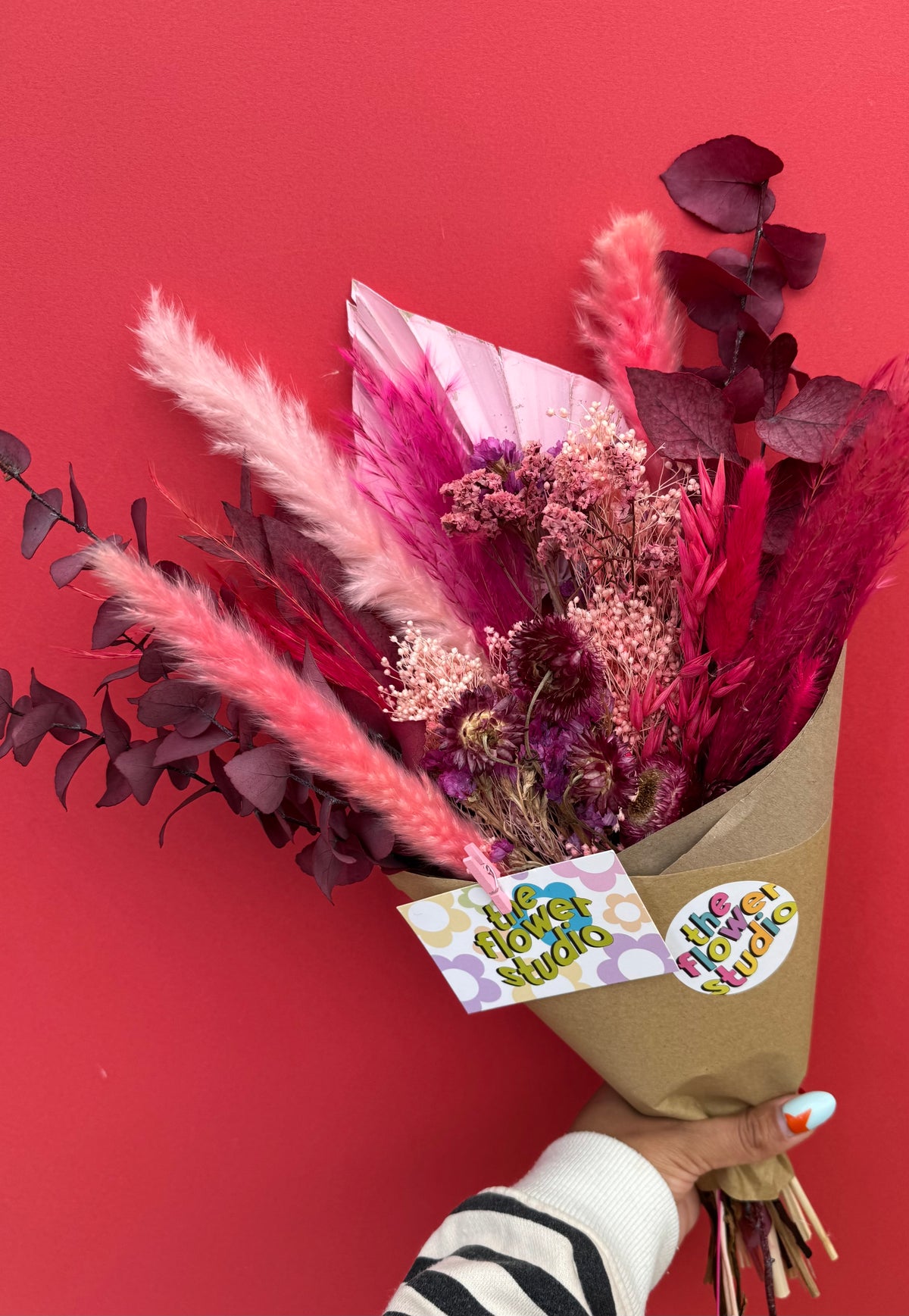 A vibrant dried flower bouquet in pink, magenta, and burgundy tones, wrapped in brown kraft paper with a 'Happy Flower Studio' tag, held by a hand against a solid red background.
