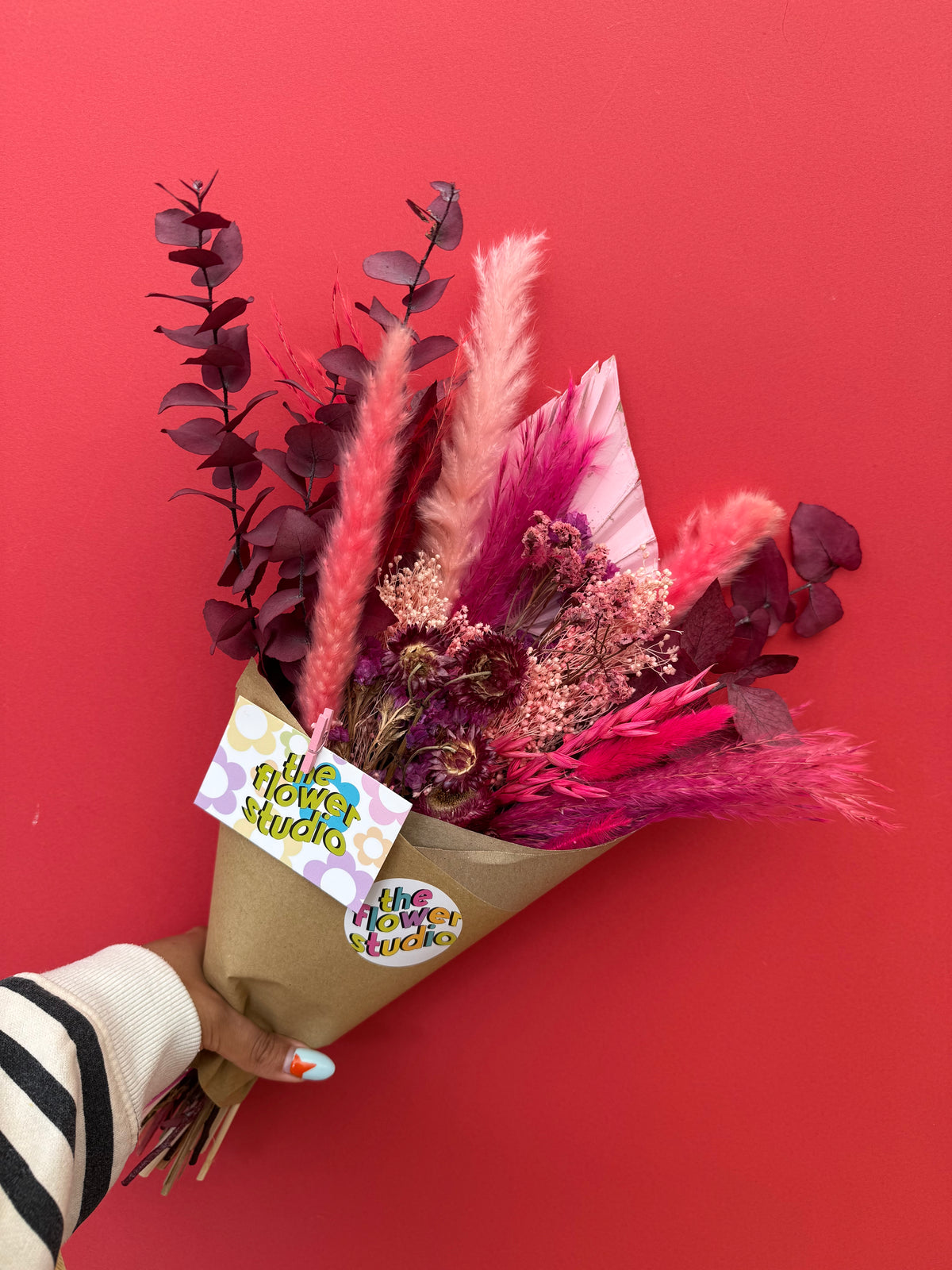 A hand holding a dried flower bouquet with pink and burgundy botanicals wrapped in brown kraft paper, featuring a 'the flower studio' tag, set against a solid red background.