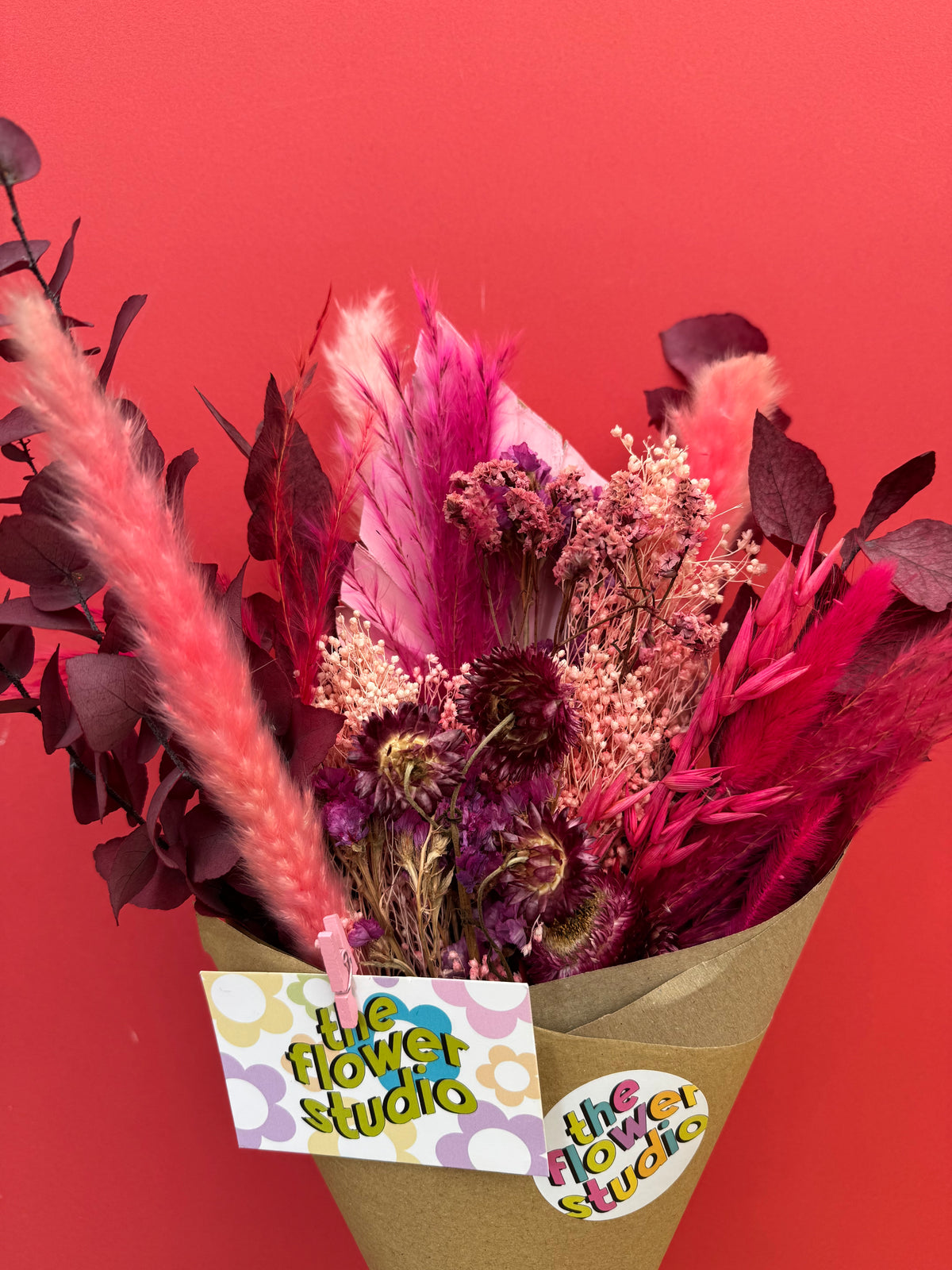 A dried flower bouquet in a brown paper cone with a 'the flower studio' tag, set against a solid red background.