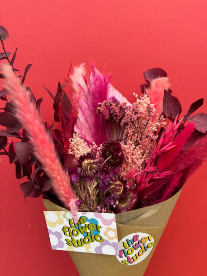 A dried flower bouquet in a brown paper cone with a 'the flower studio' tag, set against a solid red background.