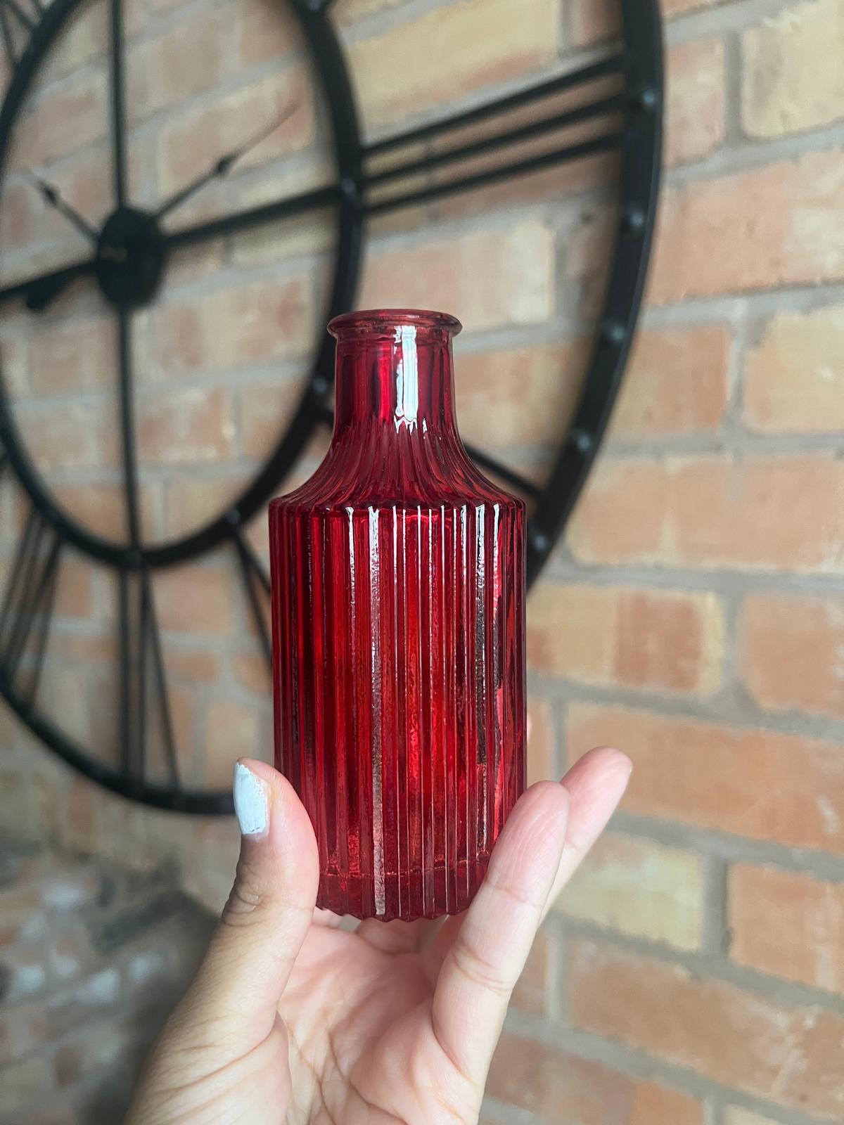Red glass bottle held by a hand in front of a brick wall with a decorative clock.