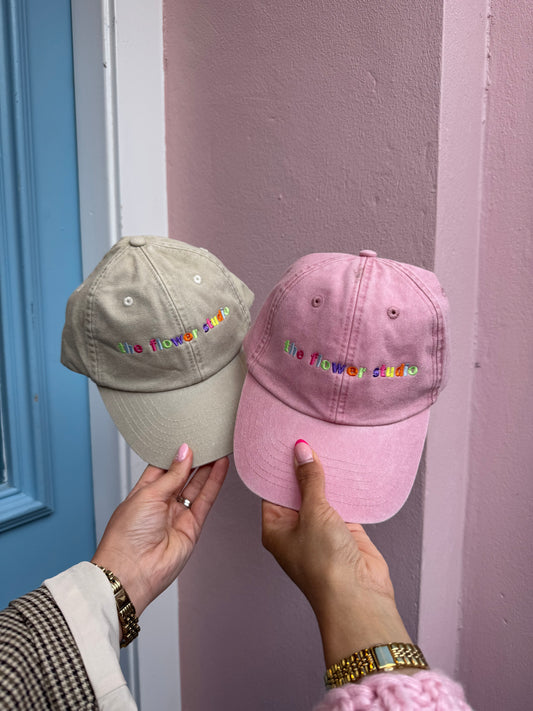 Two baseball caps, one pink and one beige, held by a person against a door and wall background.