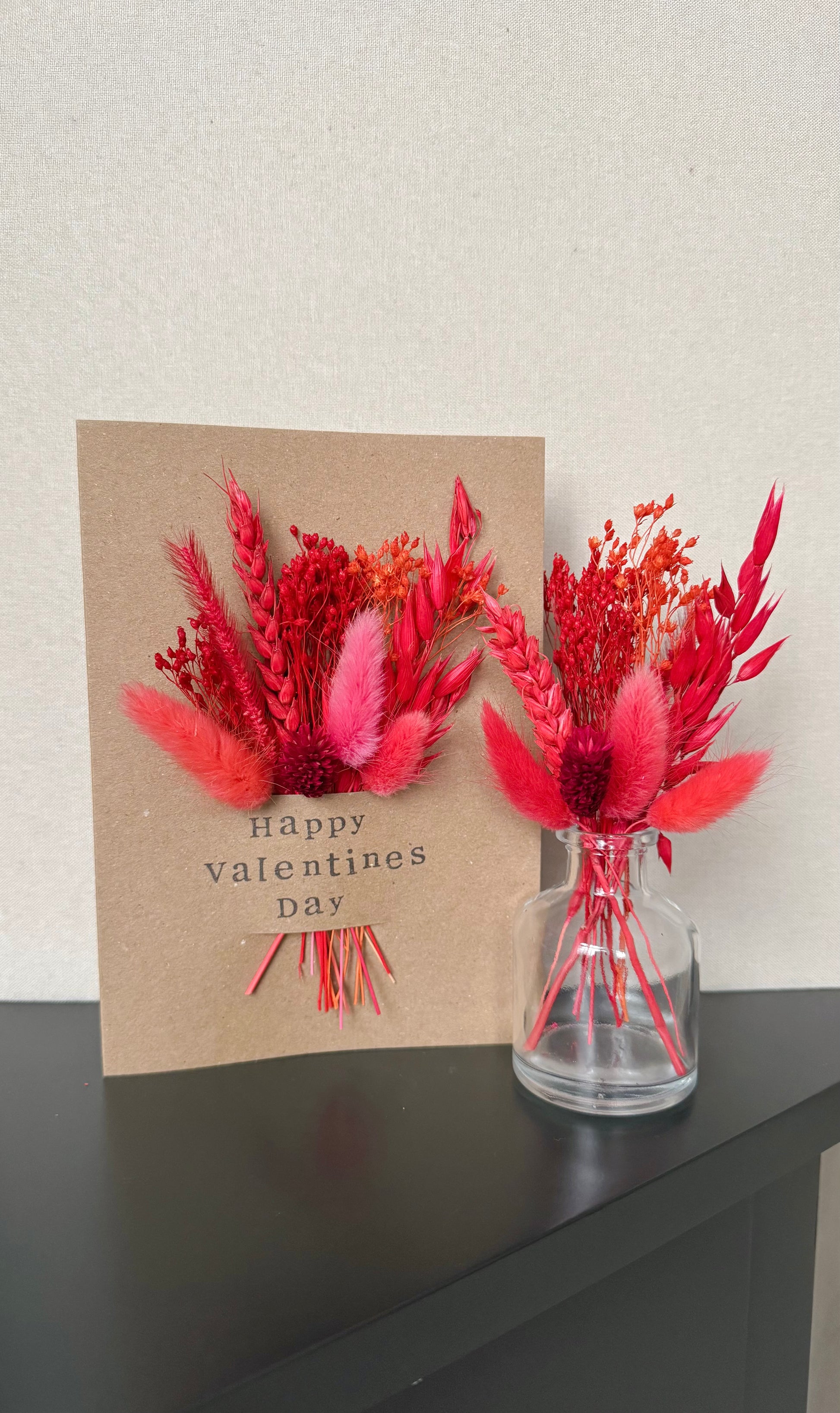Valentine's Day card with red floral arrangement and a glass vase on a dark surface.