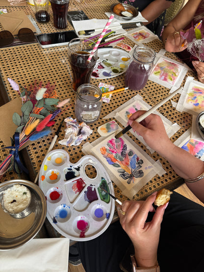A top-down view of a group of people painting together on a woven table. A hand holds a paintbrush over a canvas with colorful painted feathers, surrounded by paint palettes, jars of water and paint, and paintbrushes.