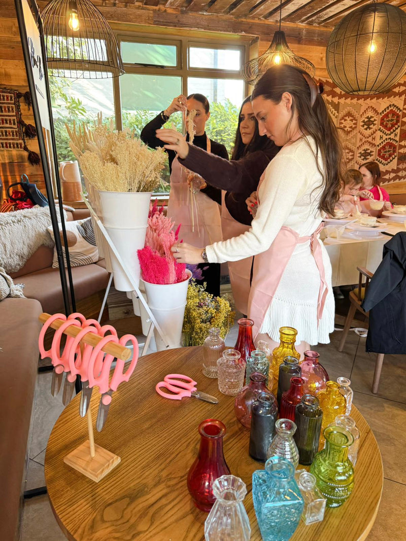 Woman in a white apron working with colorful bottles and tools in a room with large windows and wooden furniture.