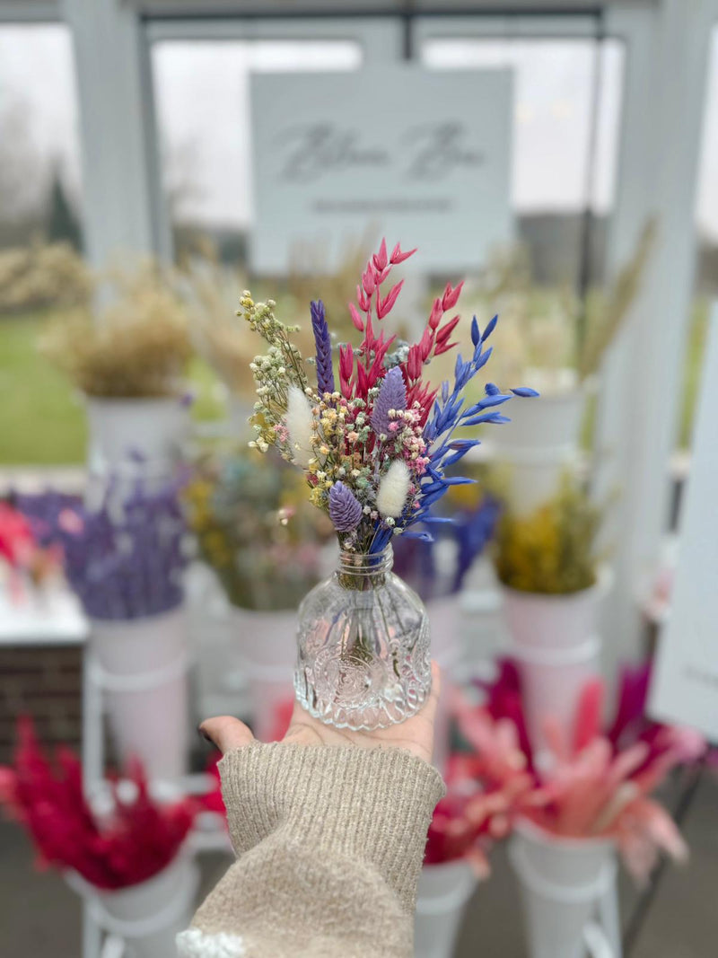 Hand holding a small bouquet of colorful flowers in a clear bottle with a blurred floral shop background.