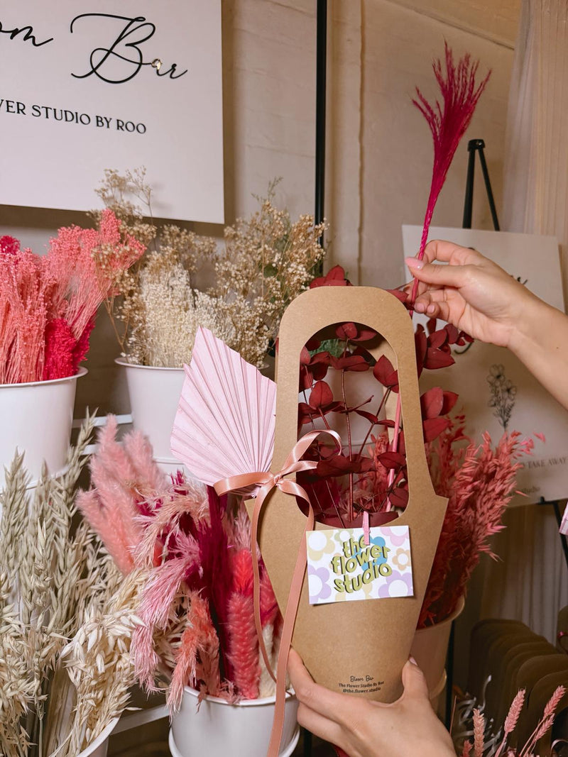 Person holding a dried flower arrangement in a wooden box with 'The Flower Studio' label, surrounded by more floral arrangements.