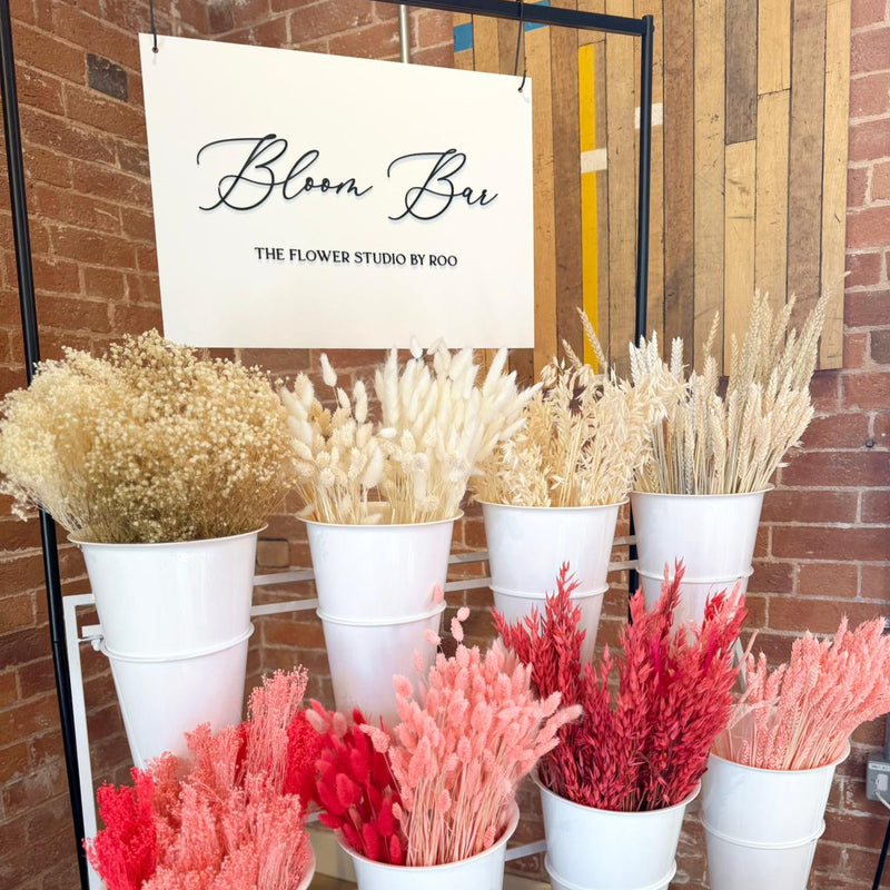 Display of dried flower arrangements in white buckets with 'Bloom Bar' sign.