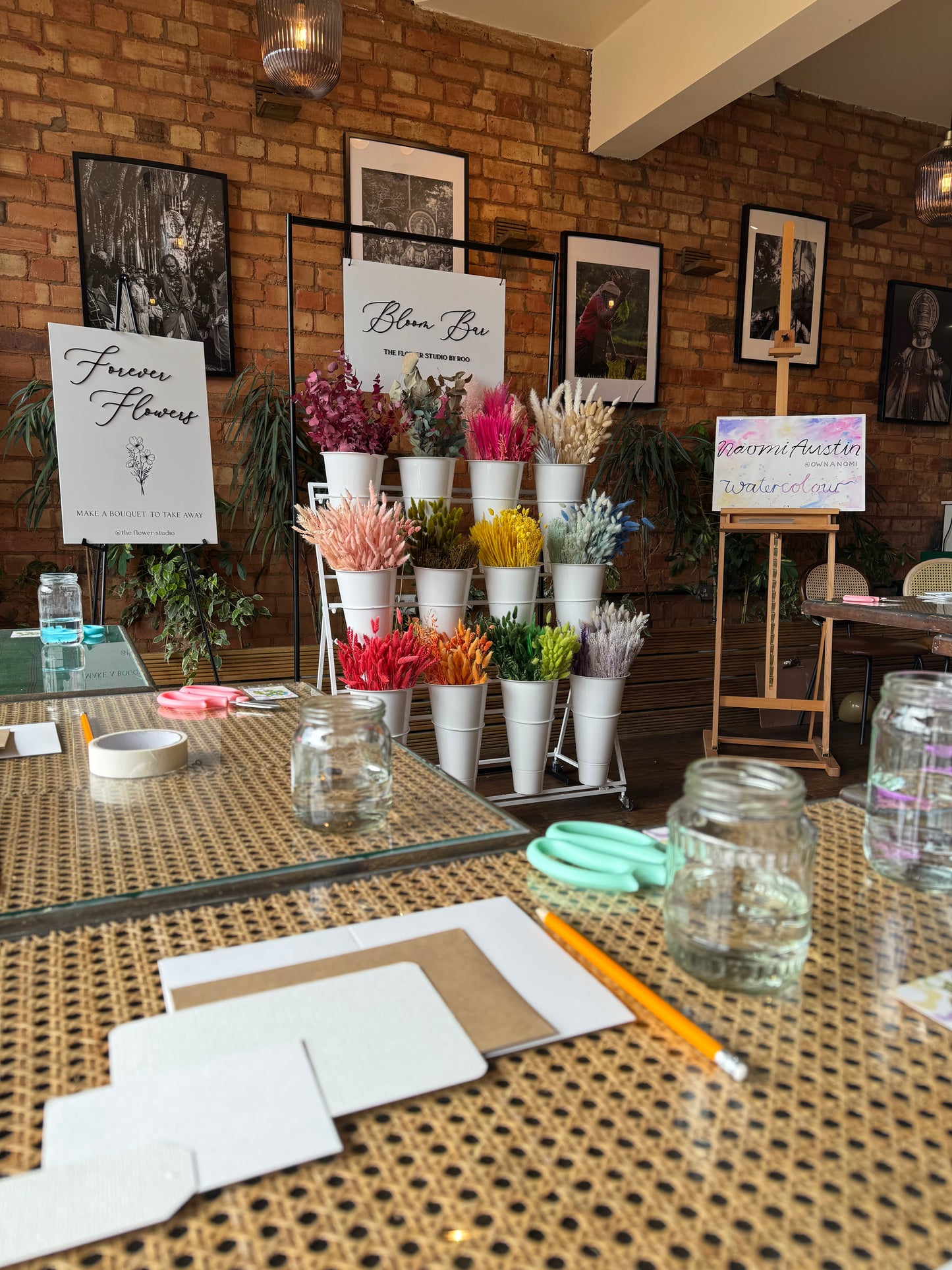 A retail display of colorful paper flowers arranged in white cups on a metal stand, set against a brick wall background with framed art and a wicker table in the foreground.